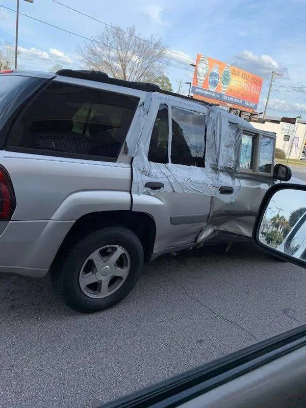 Auto s poškodenými dverami, provizórne opravené páskou na ceste v meste, reklama v pozadí.
