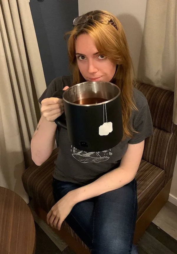 Žena na gauči drží veľký hrnček s čajom, so závesmi v pozadí.