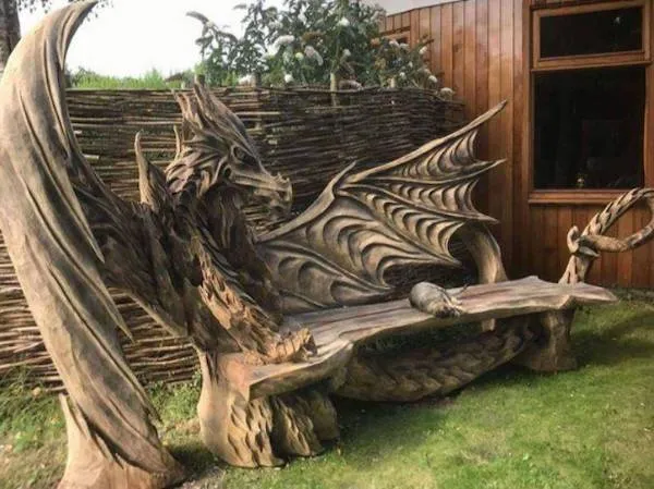 Drevená lavička v tvare draka s detailnými krídlami a hlavou pri dome na zelenej tráve.