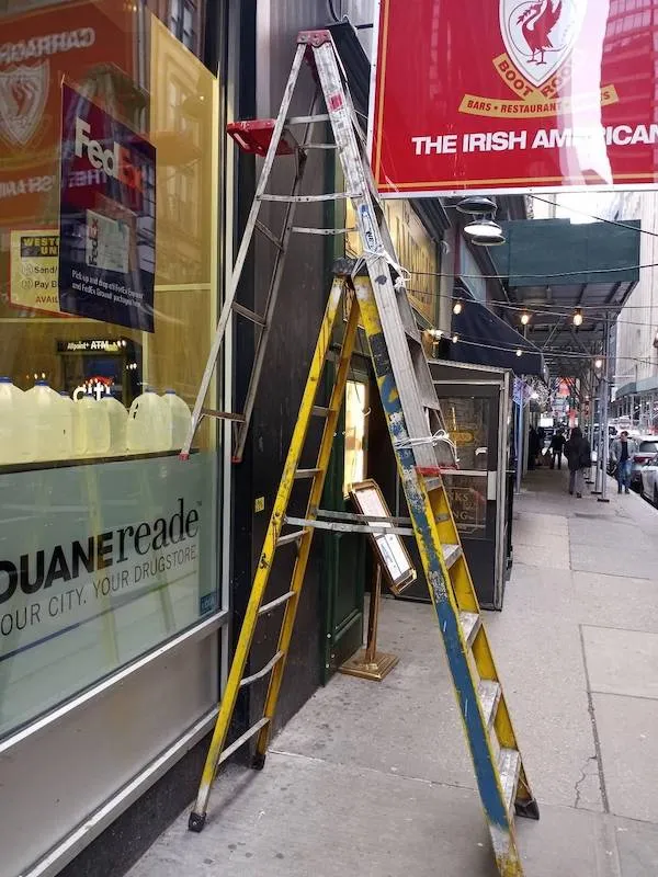 Dve rebríky opreté o stenu v meste, vedľa prevádzky Duane Reade a írskou reštauráciou na rušnej ulici.