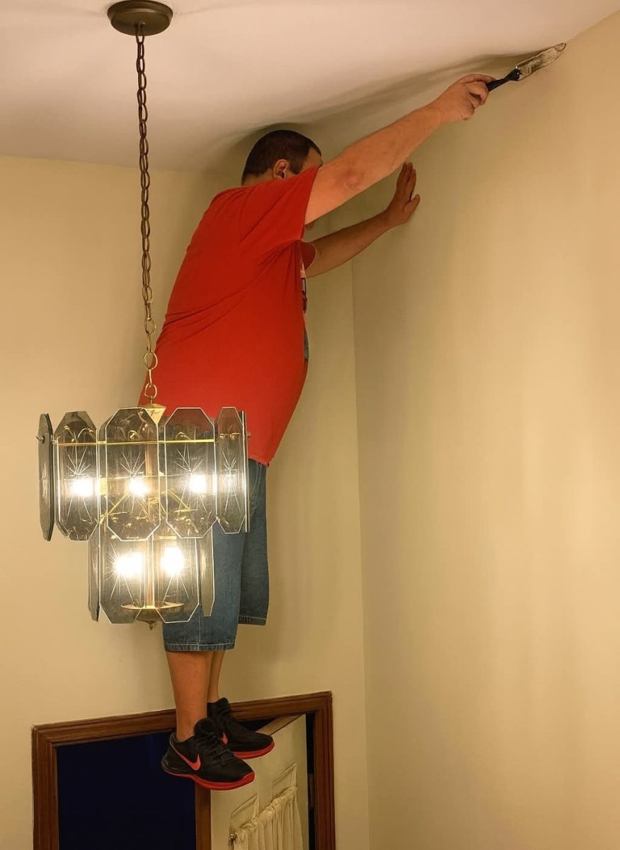 Osoba na dverách natiera strop, oblečená v červenom tričku a rifľových kraťasoch, vedľa svietidla.