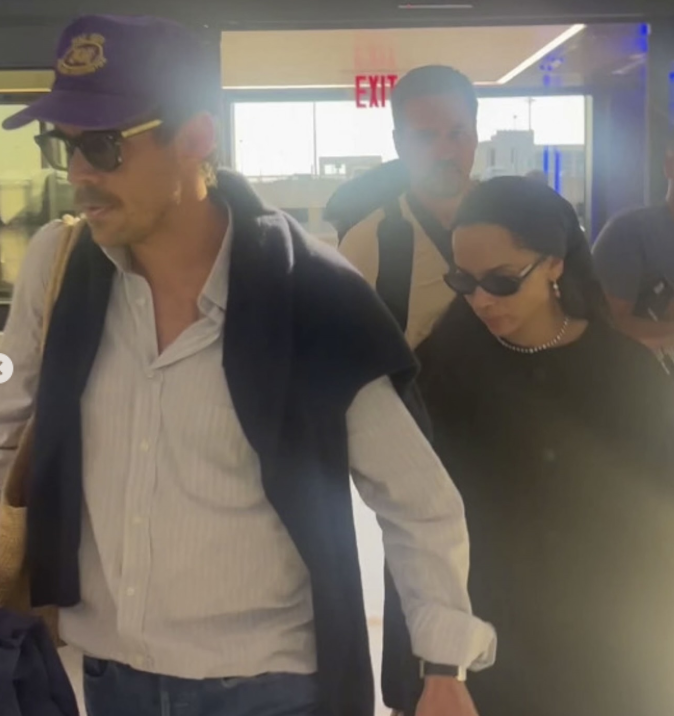 Pár kráča rušným letiskom, obaja nosia slnečné okuliare a sú oblečení v ležérnom štýle.