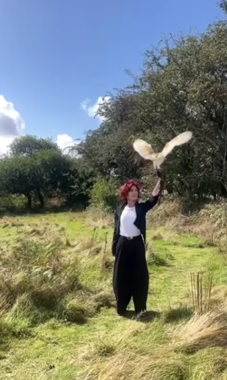 Muž stojí v tráve, dvíha ruku s rukavicou a vypúšťa sovu v prírodnom prostredí pod modrou oblohou.