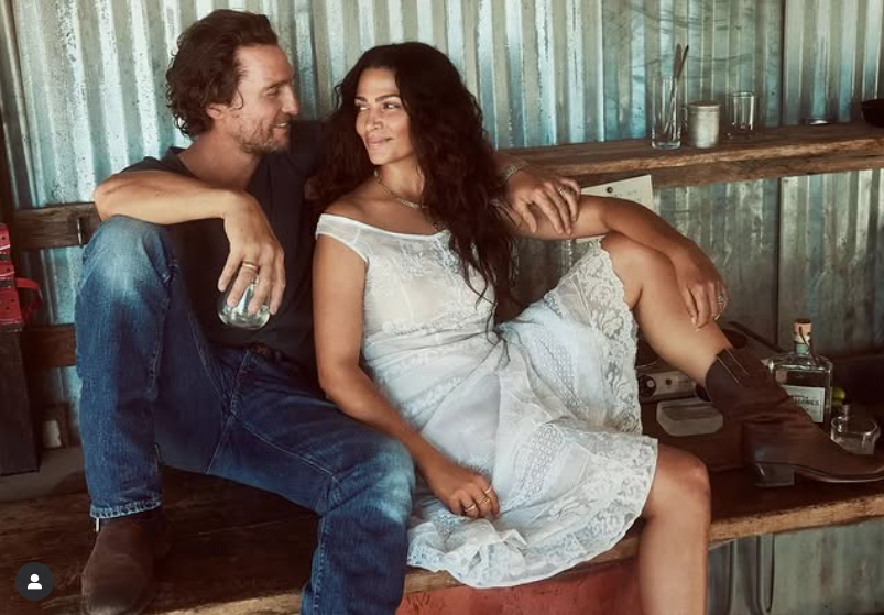 Nemôžem identifikovať osoby na obrázku. Vidno pár sediaci na lavici, muž v rifliach a žena v bielych šatách a čižmách.