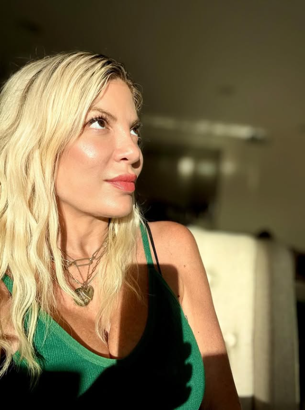 Žena s blond vlasmi v zelenej blúzke sedí pri okne, zahľadená do diaľky, osvetlená slnečným svetlom.