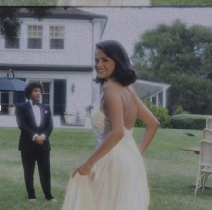 Žena v bielych šatách na záhrade s usmievajúcim sa mužom v smokingu v pozadí pred domom.