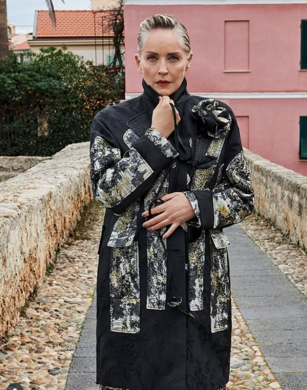 Elegantná dáma v štruktúrovanom kabáte na kamennej ceste pred ružovým domom.