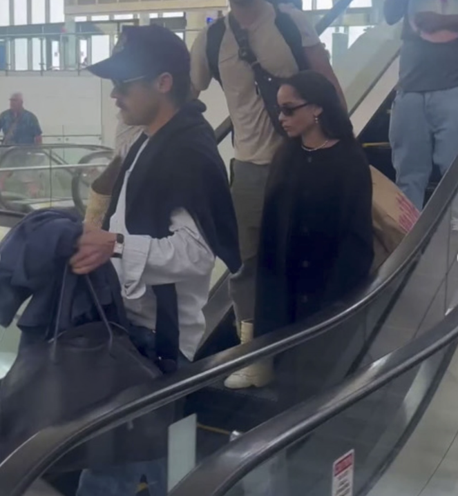 Ľudia na eskalátore v nákupnom centre, každý s batožinou a odevom pripraveným na cestovanie.