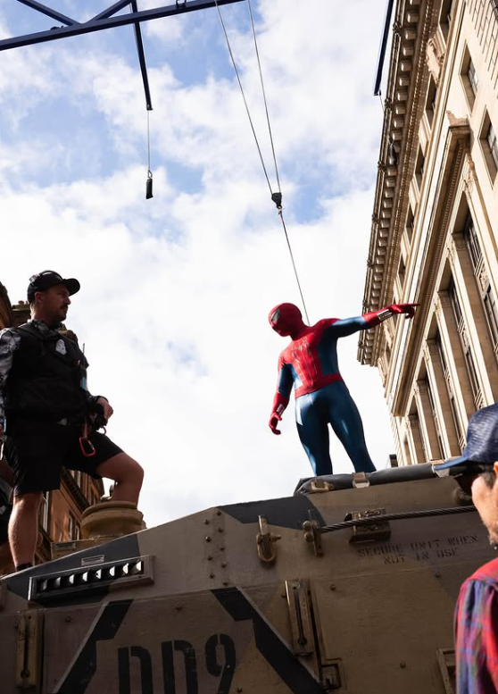 Muž v kostýme pavúčieho hrdinu na tanku počas natáčania v meste, okolité budovy a modrá obloha v pozadí.