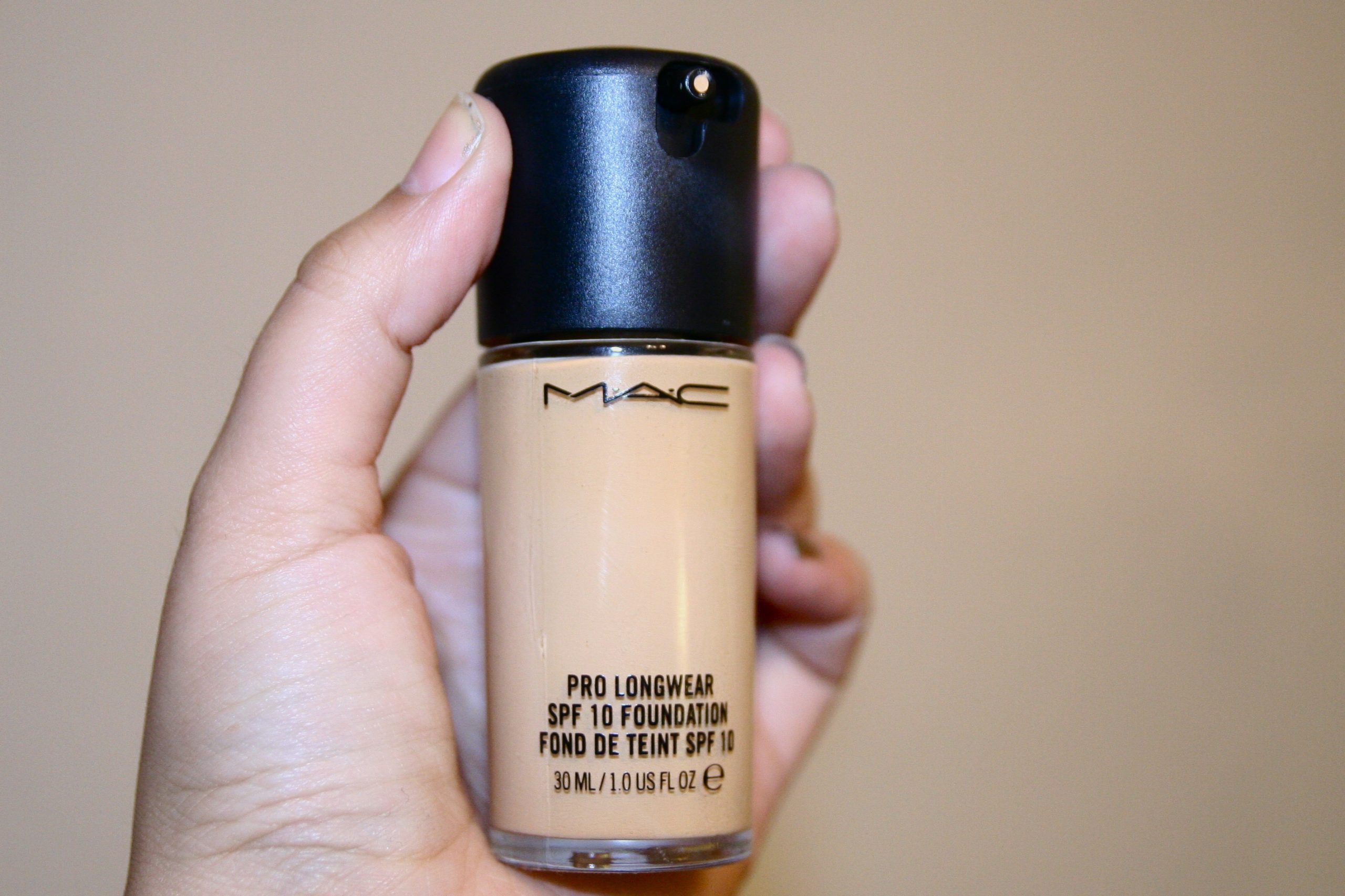 Fľaštička make-upu s čiernym uzáverom a logom MAC, držiaca ho ľudská ruka.