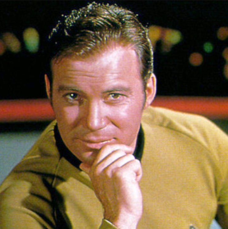 Herec v žltej uniforme na palube vesmírnej lode, zameraný pohľad a ruka pri tvári.