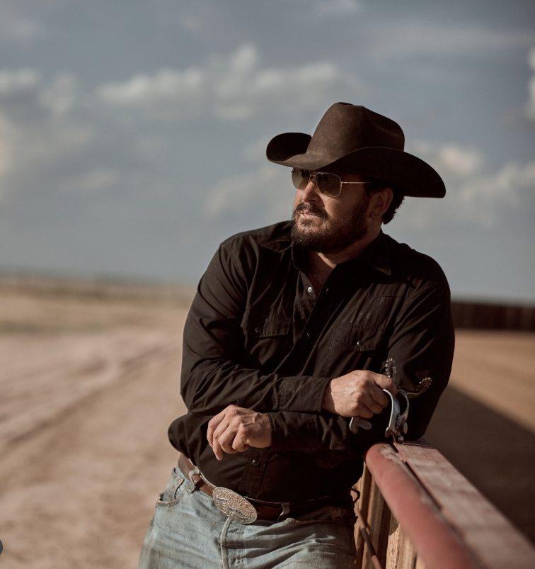 Muž v cowboyskom klobúku a slnečných okuliaroch stojí opretý o drevené zábradlie na vidieckej ceste.