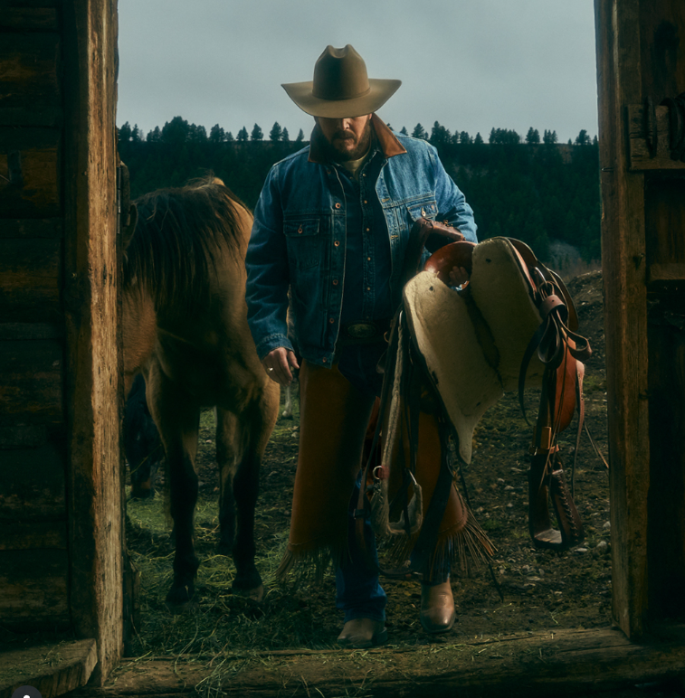 Kovboj s klobúkom vedľa koňa drží sedlo pri drevenom prístrešku, pozadie tvoria stromy a obloha.