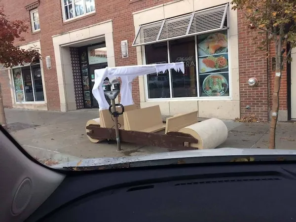 Auto v štýle Flinstonovcov zaparkované pri chodníku, pripevnené k parkovaciemu stojanu, vedľa budovy s výlohou.