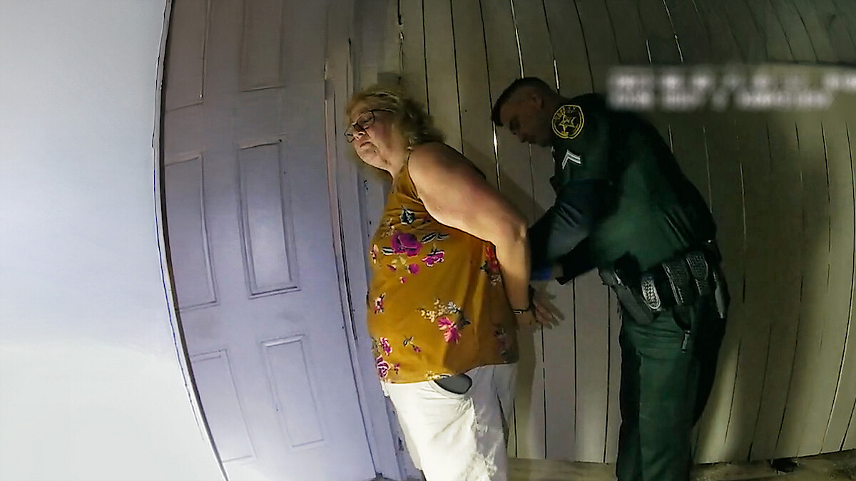 Neidentifikovaná žena zatýkaná policajtom v interiéri, ruky má zopnuté za chrbtom.