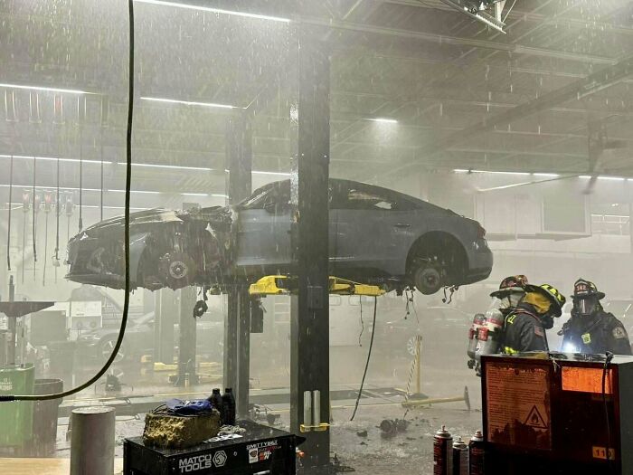 Auto na zdviháku v garáži po požiari, zasahujú hasiči v ochranných oblekoch a helmy.