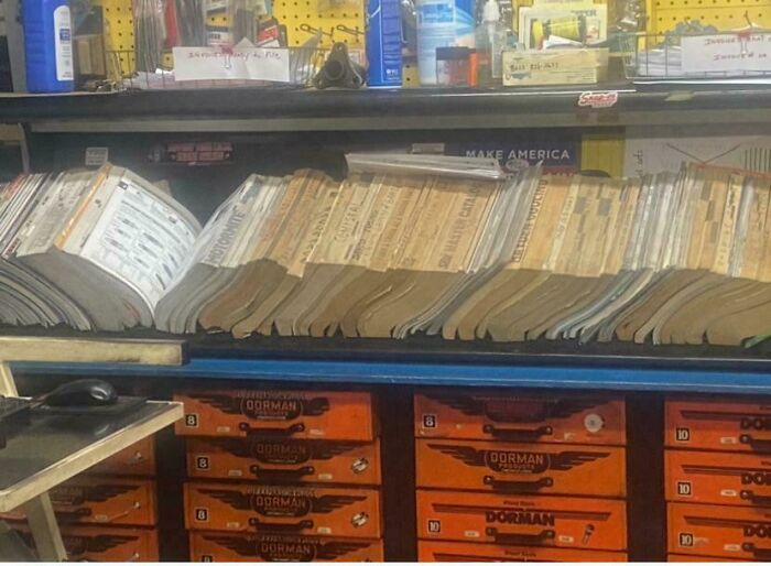 Regály s manuálmi a náradím v dielni, chaos medzi knihami a oranžovými zásuvkami na uskladnenie.