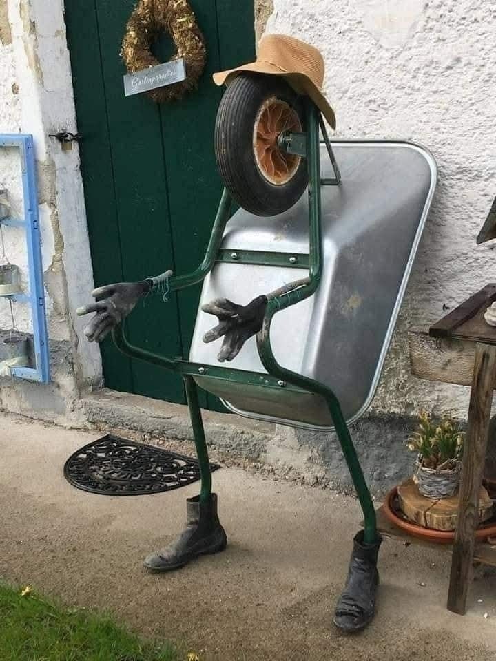 Záhradná dekorácia zo stavebného fúrika s klobúkom, rukavicami a topánkami pri dome.