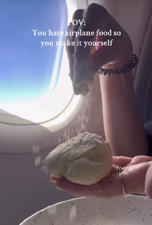 Osoba pripravuje cesto v lietadle pri okne, sype múku na guľu cesta, cez okno vidieť modré nebo.