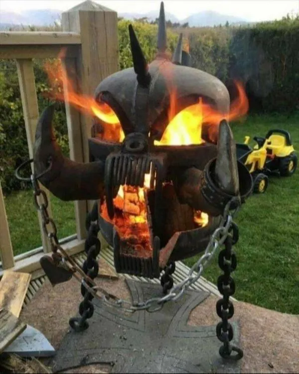 Kovová socha hlavy démona s ohňom a reťazami na dvore, v pozadí hračkárske autíčka.