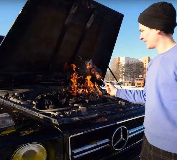 Osoba griluje na otvorenej kapote auta značky Mercedes, improvizovaný gril s ohňom vonku.