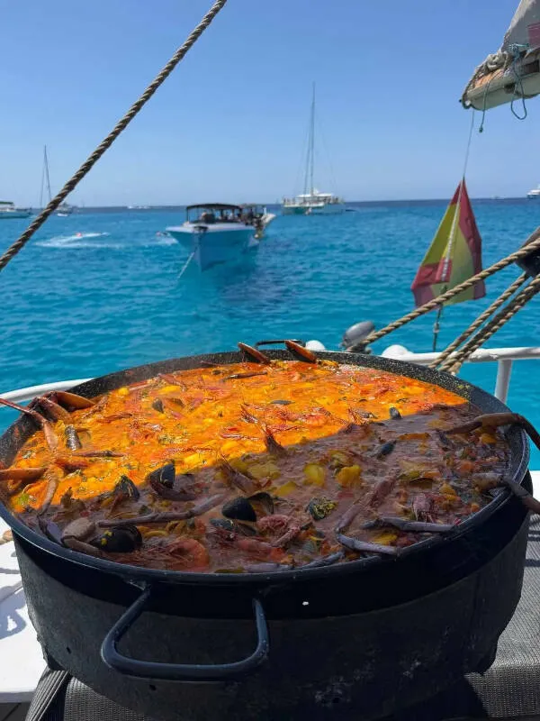 Paella s morskými plodmi na lodi, modré more a plachetnice v pozadí, slnečný deň na mori.