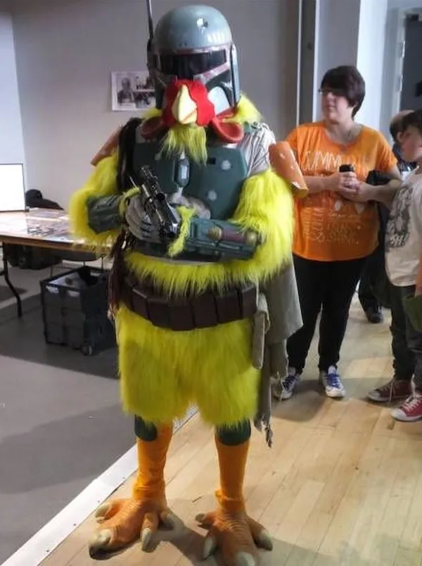 Maskot v kostýme kuraťa a sci-fi brnení s ľuďmi na pozadí v interiéri na podujatí.