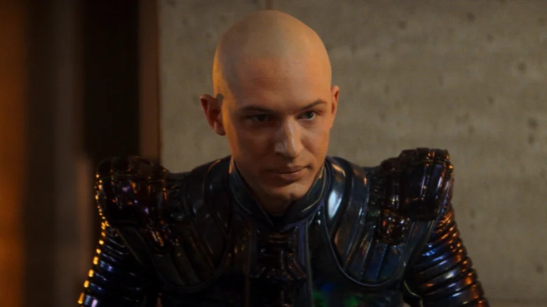 Postava v sci-fi kostýme s výrazným brnením a holou hlavou, stojí na čelnej strane v tlmenom osvetlení.