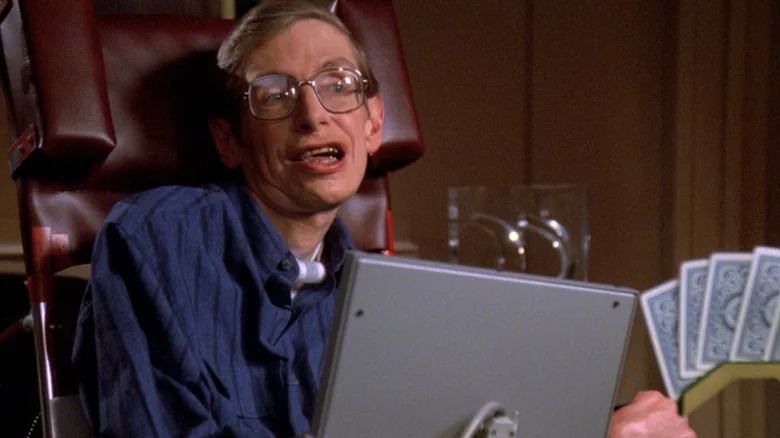 Osoba v modrej košeli na vozíku s kartami v pozadí, sedí pri stole s počítačovou obrazovkou.