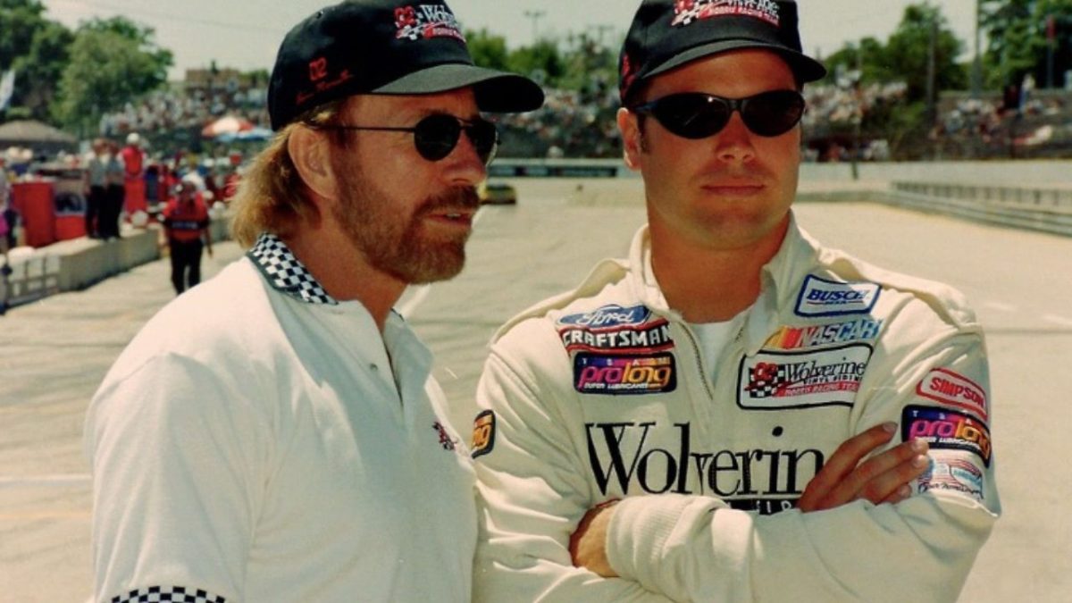 Dve osoby v športových okuliaroch na automobilových pretekoch v diskusii, nosia čiapky a pretekárske oblečenie.