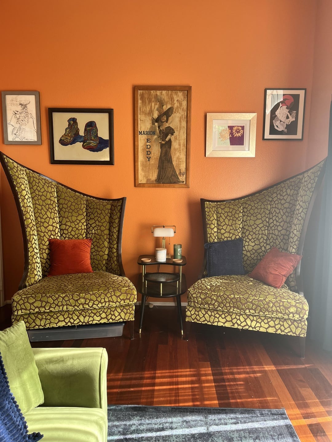 Dve zelené kreslá s oranžovými vankúšmi, obklopené obrazmi na oranžovej stene s moderným dizajnom interiéru.