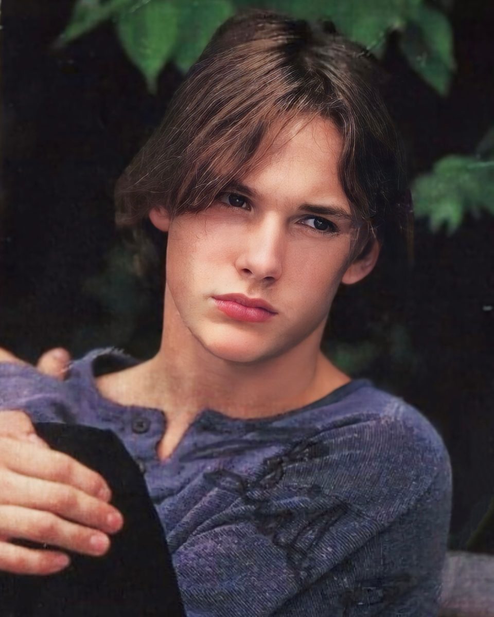 Osoba s dlhšími vlasmi v modrom svetri sedí na tmavom pozadí zelených listov.