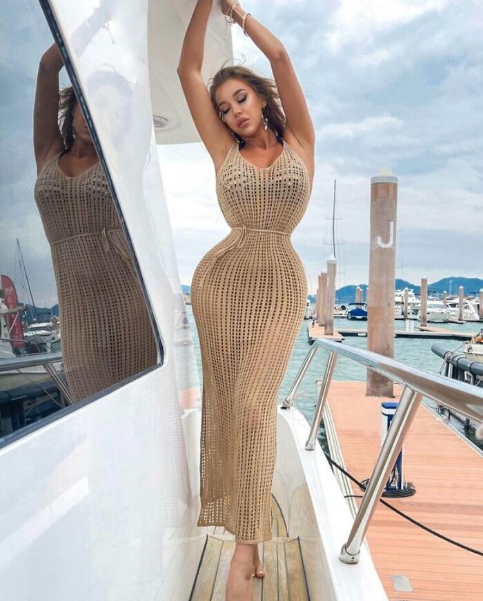 Žena v pletených šatách pózuje na jachte s prístavom v pozadí.