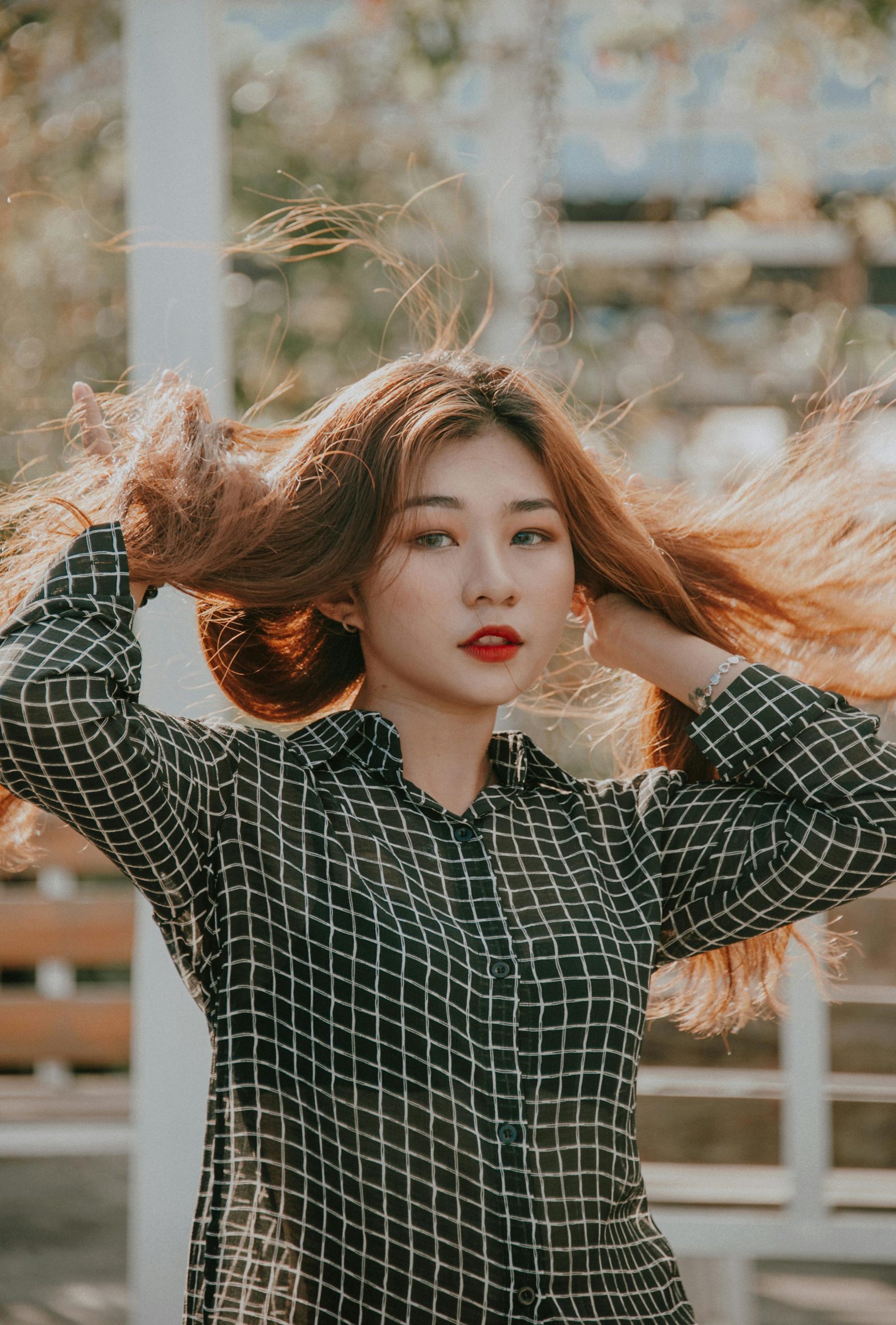 Žena s dlhými vlasmi v čiernej kockovanej blúzke vo vetre, prírodné pozadie s jemným bokeh efektom.