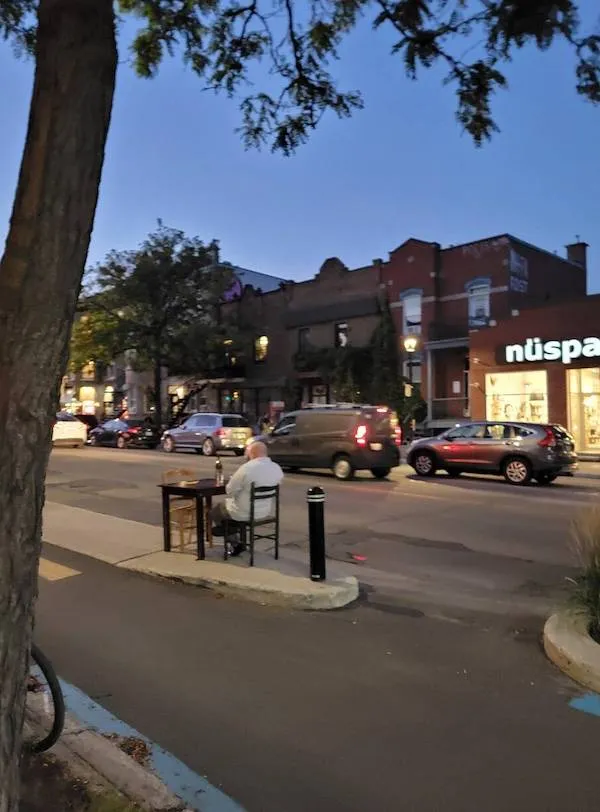 Starší pán sedí pri stole blízko cesty v mestskom prostredí, za súmraku, okolo jazdia autá.