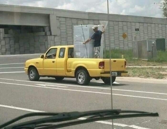 Žlté auto preváža sklený panel, muž ho zabezpečuje na korbe počas jazdy na otvorenej ceste.