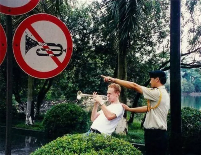 Muž hrá na trúbku pred zákazovou značkou, zatiaľ čo ho zastavuje ochranka v parku.