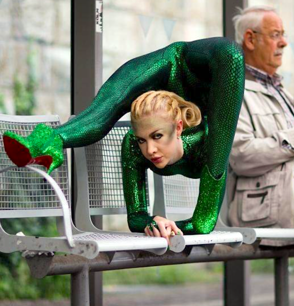 Žena v zelenom akrobatickom obleku predvádza flexibilitu na lavičke, sediaci muž ju ignoruje.