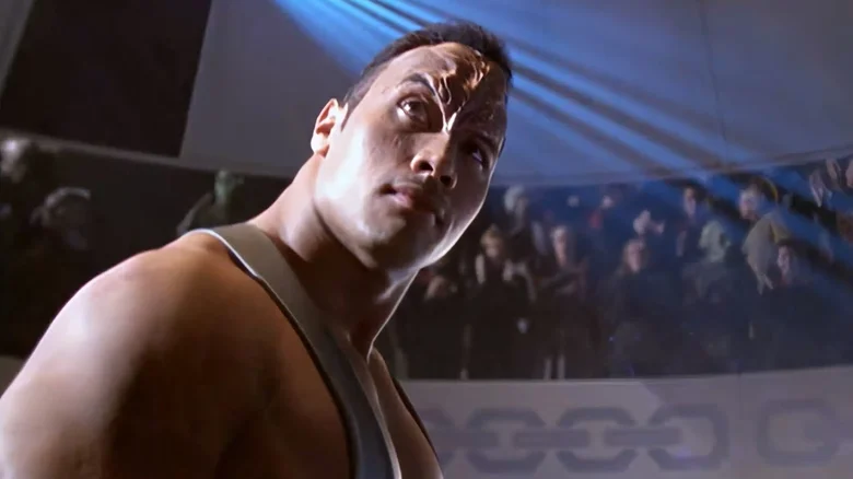 Osoba v bojovej scéne s intenzívnym výrazom, futuristické osvetlenie a diváci v pozadí, atmosféra napätia.