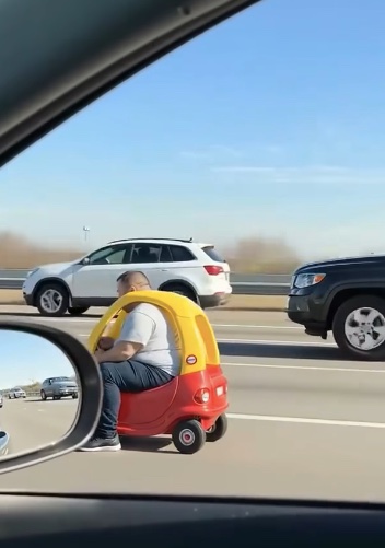 Dospelý muž na diaľnici sedí v malom plastovom autíčku vedľa premávky.