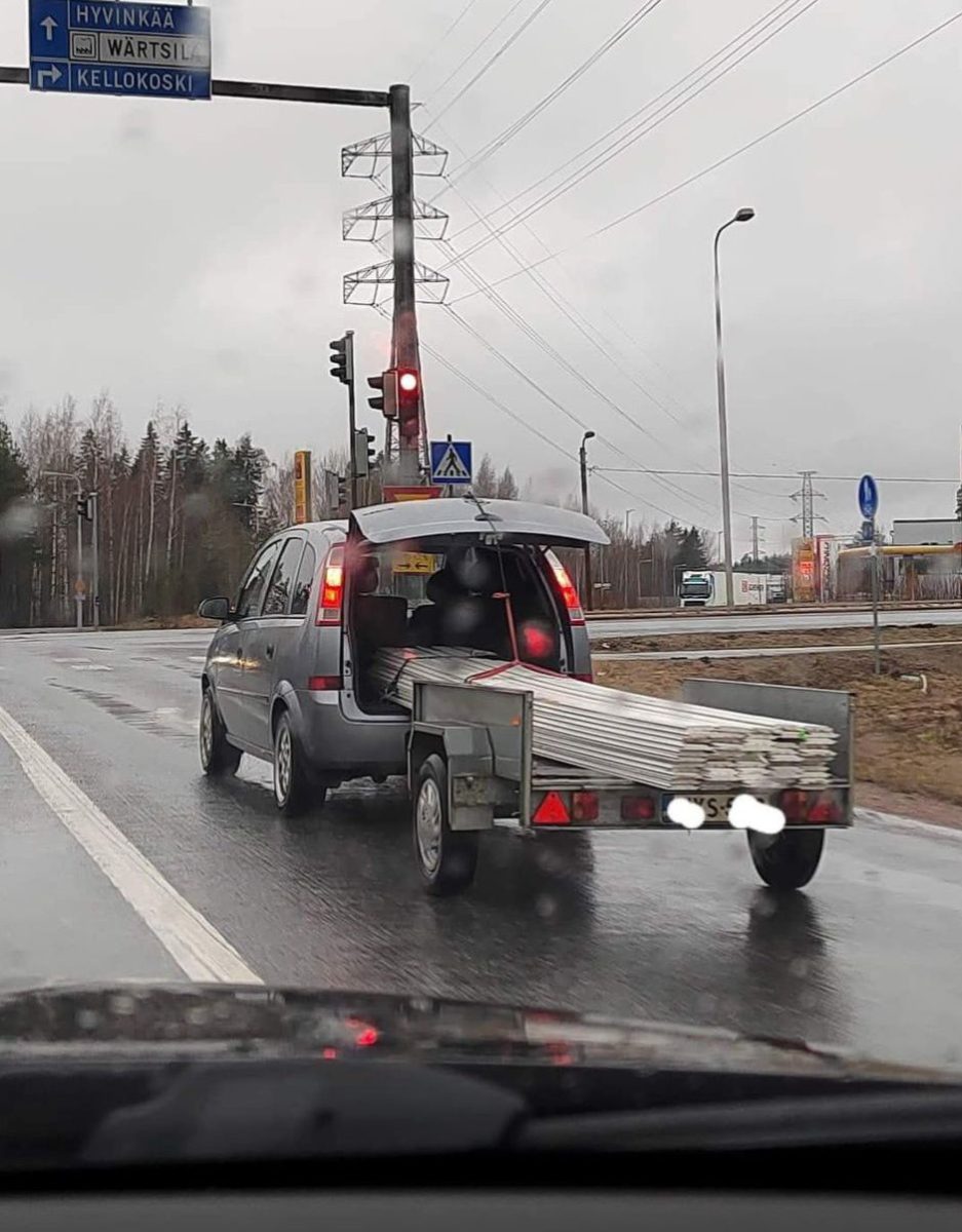Auto s otvoreným kufrom a prívesným vozíkom nesie dlhé kovové tyče na mokrej ceste pod oblačným nebom.