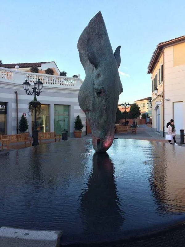 Socha konskej hlavy nad vodnou hladinou v mestskom námestí pod modrou oblohou.