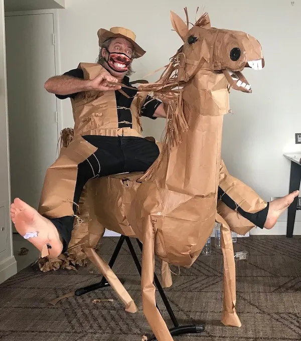 Muž v zábavnom kostýme na papierovom koni s úsmevom v izbe.
