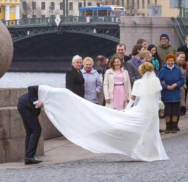 Svadobná fotka pri moste: nevesta so závojom a ženích vytvárajú veselú atmosféru; sleduje ich skupina svadobčanov.