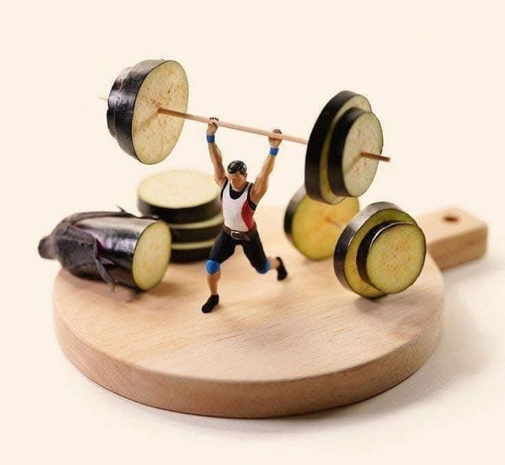 Miniatúrny kulturista dvíha činku z plátkov baklažánu na drevenej doske. Kreatívna kuchynská kompozícia.