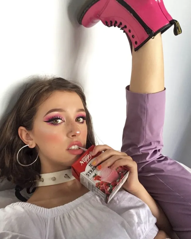 Osoba s extravagantným líčením a fuksiovými čižmami pije jahodový džús, nohu má zdvihnutú vysoko do vzduchu.