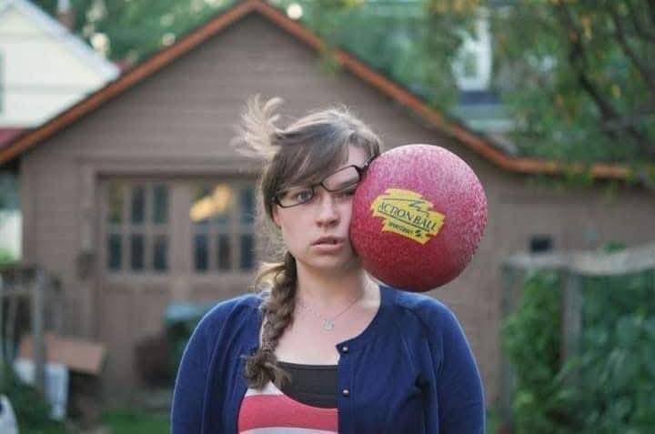 Mladá žena s okuliarmi zasiahnutá loptou do tváre v záhrade.