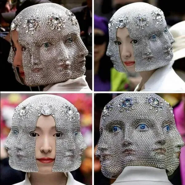 Osoba s extravagantnou pokrývkou hlavy zdobenou tvárami a trblietavými krištáľmi, pripomínajúca umelecký módny doplnok.