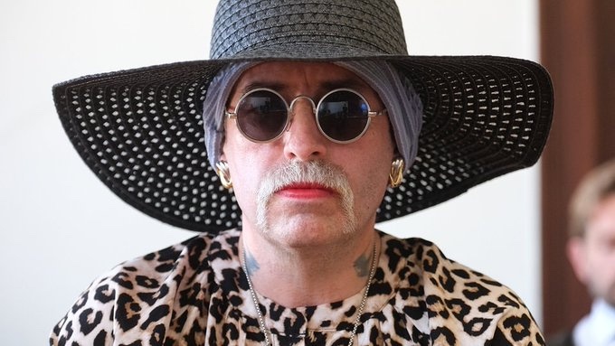 Osoba v leopardích šatách s veľkým klobúkom a slnečnými okuliarmi, výrazný štýl.