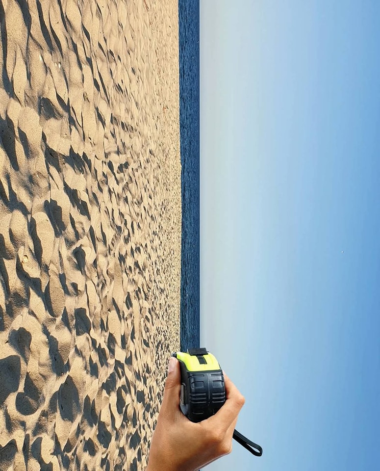 Ruka držiaca merací pás na piesočnatej pláži s výhľadom na more a modrú oblohu.